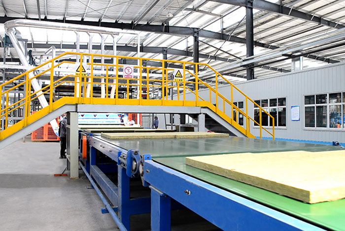 Stone wool boards moving along a factory conveyor line beneath overhead piping and a steel-frame roof.